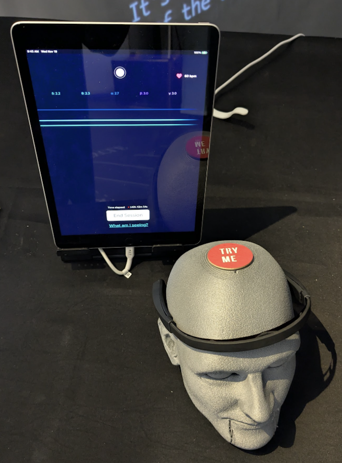 The BrainCo's Focus 1 is an EEG device that fits across the wearer's forehead and above their ears. This device is black, and in this image, it is displayed on a grey mannequin head. Behind it is an iPad showing the EEG software readout.